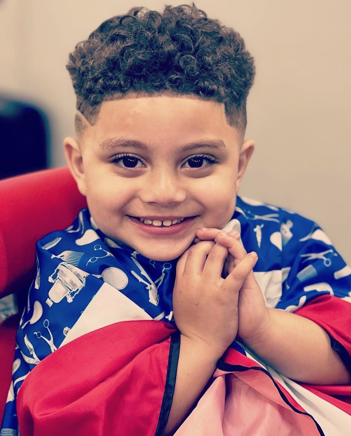 Niño sonriente con corte rizado en capa de barbería — X Express Barbershop